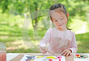 Happy asian child girl sitting at a table in a summer garden painting with paintbrush, Education art concept