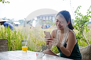 Happy Asian Chinese woman on her 20s or 30s smiling having fun using internet on mobile phone drinking orange juice sitting outdoo