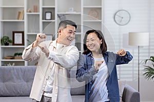 Happy Asian couple man and woman dancing together at home in daytime in living room, happy young family celebrating