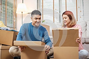 Happy asian couple open cardboard boxes while moving to new home at sofa in living room.unpacking for new house