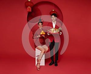 Happy asian couple in red casual attire holding fresh fruit and orange bless for Chinese new year day on red room decoration