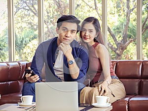 Happy Asian couple sitting on couch in living room with computer laptop and coffee cup on table smiling and looing to camera