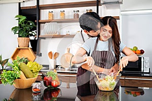 Happy Asian couple wearing aprons, tossing the vegetarian salad with wooden spatulas. Preparing a healthy salad.