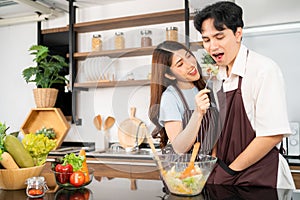 Happy Asian couple wearing aprons, a Woman feeding the vegetarian salad to a man for taste. Cooking a healthy salad.