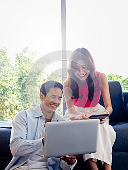 Happy Asian couple, young woman sit on sofa and man on floor looking and using laptop computer.