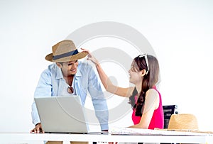 Happy Asian couple, young woman put hat on man head with smile while using laptop computer on desk together.