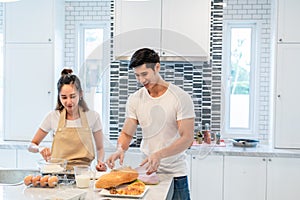 Happy Asian couples cooking and baking cake together in kitchen room. Man and woman looking to tablet follow recipe step at home.