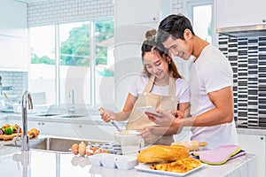 Happy Asian couples cooking and baking cake together in kitchen room. Man and woman looking to tablet follow recipe step at home.