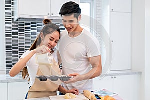 Happy Asian couples cooking and baking cake together in kitchen room. Man and woman looking to tablet follow recipe step at home.