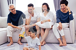Happy Asian extended family sitting on sofa together and watching little child playing toy on the floor with happiness.