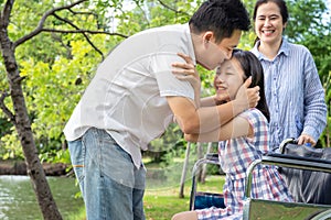 Happy asian family,beautiful sick daughter in wheelchair while her father hugging,kissing on the forehead her and mother smiling