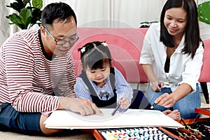 Happy Asian family. Chubby little girl daughter drawing together with father and mother in living room. Parents spend time with