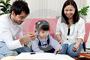 Happy Asian family. Chubby little girl daughter drawing together with father and mother in living room. Parents spend time with