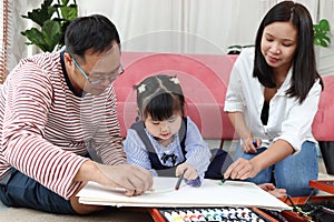 Happy Asian family. Chubby little girl daughter drawing together with father and mother in living room. Parents spend time with