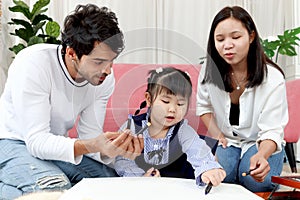 Happy Asian family. Chubby little girl daughter drawing together with father and mother in living room. Parents spend time with