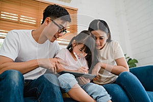 Happy Asian family dad, mom and daughter using computer tablet technology sitting sofa in living room at house. Self-isolation,