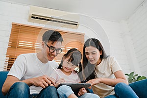 Happy Asian family dad, mom and daughter using computer tablet technology sitting sofa in living room at house. Self-isolation,