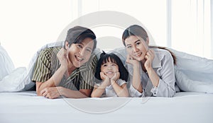 Happy Asian family with a daughter are lying and Putting hand on chin under the blanket on bed in the bedroom