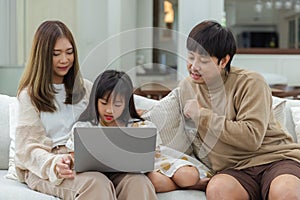 Happy asian family father mother and child daughter having fun using laptop computer sitting on couch in living room at home.