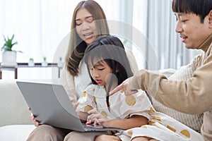 Happy asian family father mother and child daughter having fun using laptop computer sitting on couch in living room at home.