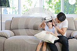 Happy Asian family, father teaching daughter to play virtual reality goggles on the sofa in the living room with happy smiling