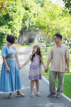 Happy Asian family holding hands and walking, enjoying leisure time with outdoor activities in a public park.