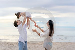 Happy Asian family holidays during joyful father, mother and daughter walking and raise daughter to sky along summer sunset sea.