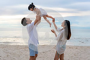 Happy Asian family holidays during joyful father, mother and daughter walking and raise daughter to sky along summer sunset sea.
