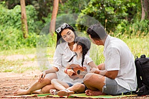 Happy asian family picnic. daugther playing ukulele with her parents Father, Mother singing song in park