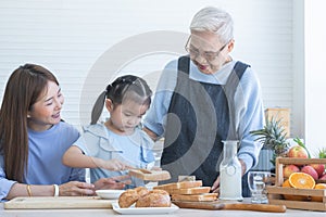 Happy Asian family preparing breakfast together. Grandmother and mother looking at child daughter girl spreading fruit jam on