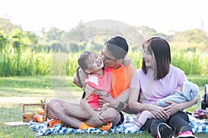 Happy Asian family in sportswear picnic at park, father kissing cute little daughter, mother smiling and holding newborn baby