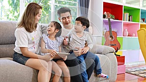 Happy Asian family teaching children son and daughter how to use tablet while sitting on grey sofa in living room with smiling fac