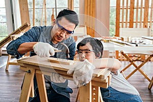 Happy Asian father and son work as a woodworker and carpenter. Father teaching his son to hammer nails on a wooden plank carefully