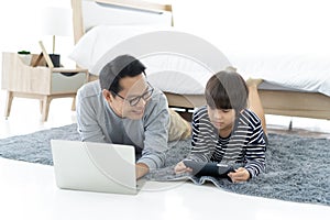 Happy Asian father using a laptop computer and his cute little son using a digital tablet while lying on the carpet in bedroom.
