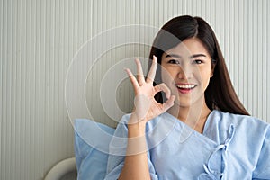 Happy Asian female patient smiling, lies on the bed, and Shows an ok symbol to show confidence in treatment. Concept of believe in