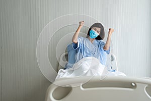 Happy Asian female patient wearing a mask, lies on the bed, and I raised an arms and was delighted for show confidence in