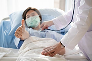 Happy Asian female patient wears a mask, lies on the bed, and raises her thumb up. When a doctor uses a stethoscope to listen to
