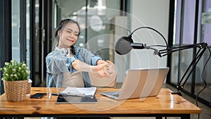 A happy Asian female radio host stretches her arms at her desk after finishing her live radio show