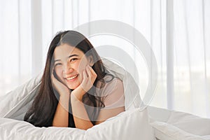 Happy Asian girl with black long hair lying on the bed with hands touching her face and giving sweet smile and giving eye contact