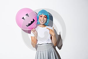 Happy asian girl celebrating halloween and wearing blue wig for party, pointing finger at balloon with scary face