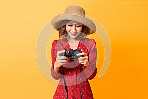 Happy Asian girl checking photos on her camera while wearing red dress and hat isolated on orange background