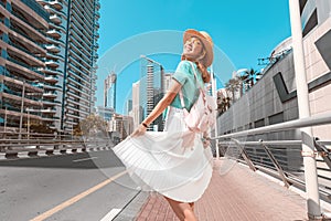 Happy Asian girl in a dress walks through a modern neighborhood with skyscrapers and office buildings. Tour and travel in Dubai