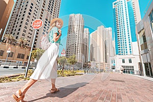 Happy Asian girl in a dress walks through a modern neighborhood with skyscrapers and office buildings. Tour and travel in Dubai