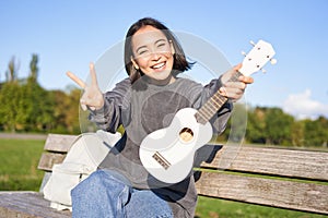 Happy asian girl shows ukulele and thumbs up, demonstrates her new musical instrument, learns how to play in park, sits