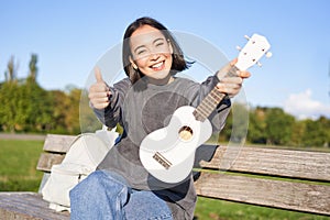 Happy asian girl shows ukulele and thumbs up, demonstrates her new musical instrument, learns how to play in park, sits