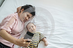 Happy Asian Grandmother and little baby boy or Grandson lying on bed taking selfie photos and doing video call with mobile phone