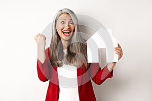 Happy asian grandmother showing paper house cutout and fist pump gesture, scream yes with joy, buying property, standing