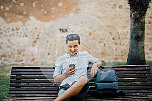 Happy asian guy using a smart phone sitting on a bench in a park