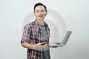 Happy asian indonesian man holding smartphone and laptop computer on isolated background