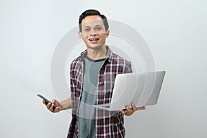 Happy asian indonesian man holding smartphone and laptop computer on isolated background